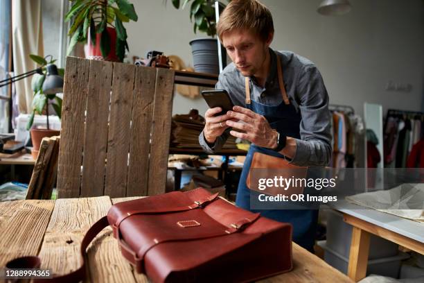 craftsman taking cell phone picture of satchel in his workshop - e commerce imagens e fotografias de stock