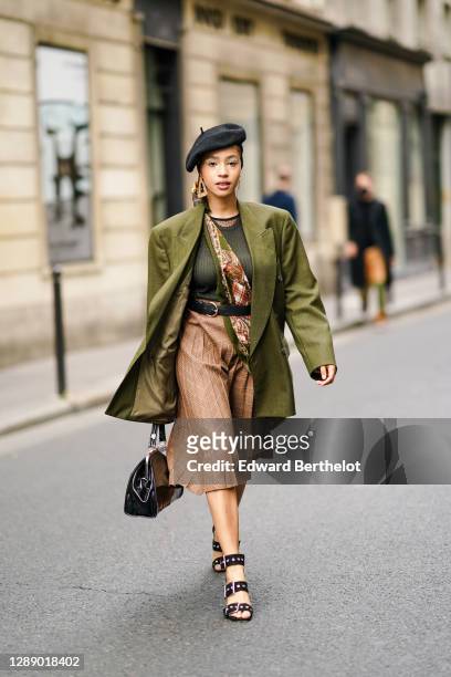 Guest wears a black beret hat, earrings, a green khaki oversized blazer jacket, a mesh top, a floral print scarf, a brown pleated checked skirt, a...
