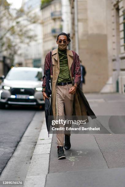 Guest wears sunglasses, a brown face mask, a red/burgundy and beige padded trench coat, a green satin turtleneck pullover, a leather belt, beige...