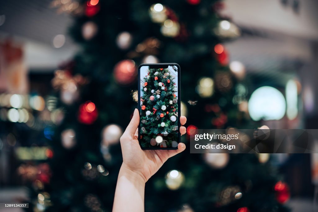 Personal perspective of woman's hand taking photo of a colourful Christmas tree with smartphone in the festive Christmas season