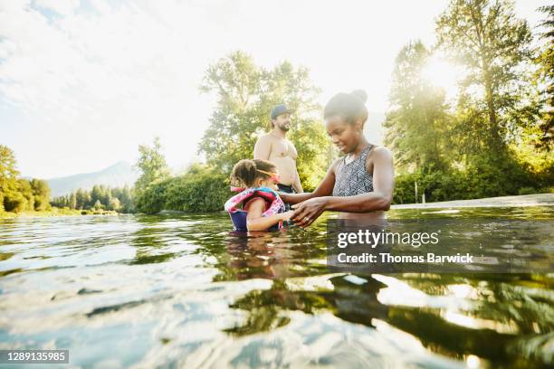 smiling mother helping young daughter learn to swim in river on summer afternoon - kids at river stock pictures, royalty-free photos & images