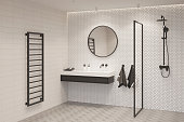 A bright bathroom with a round mirror over a black and white washbasin, with two terry towels, a glass partition, a shower, a black heated towel rail.