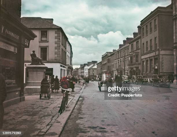 Pony cart carries churns of milk past shoppers and cars parked on Main Street in the town of Tipperary in County Tipperary, Ireland in June 1948.