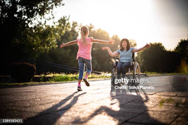 vista trasera jovencita va a abrazar a las mujeres en silla de ruedas - silla de ruedas fotografías e imágenes de stock