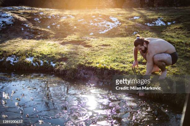 ein junger mann mit langen haaren und starkem körper, der am ufer am see steht und einen sonnigen wintertag genießt - ein mann bereitet sich darauf vor, ins eisige wasser zu kommen - eis baden stock-fotos und bilder