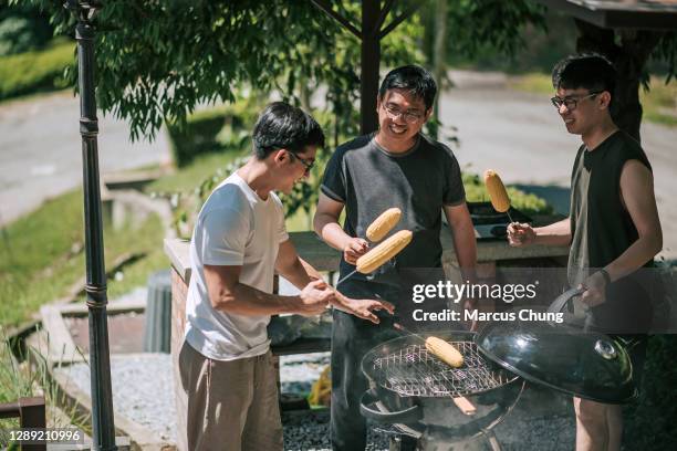 amici maschi sorridenti cinesi asiatici che grigliano insieme il mais dolce nel cortile di villa - bbq corn foto e immagini stock