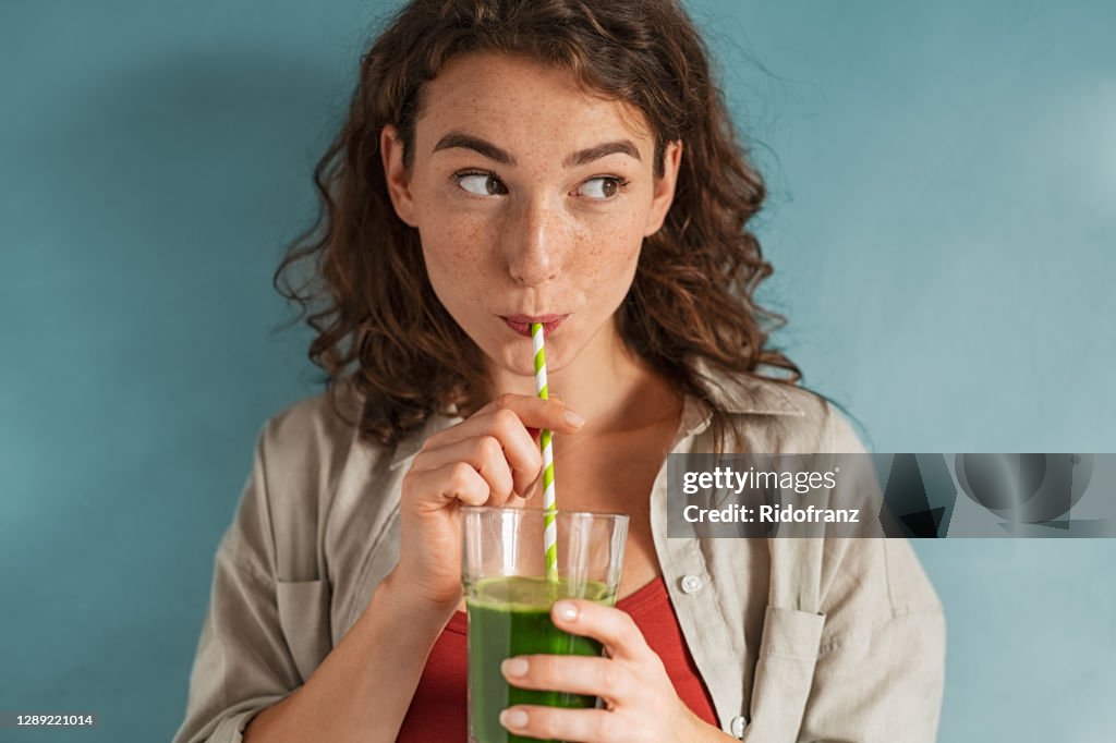 Young woman drinking detox juice with straw on blue wall