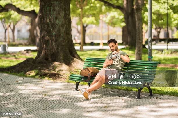 joven se relaja en un banco del parque y vertiendo té mate de un matraz - yerba mate fotografías e imágenes de stock