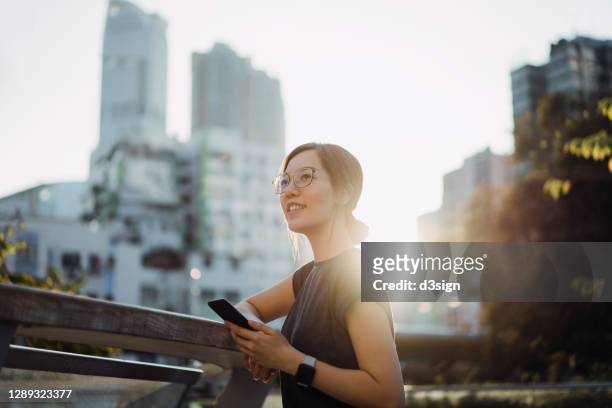 beautiful smiling young asian woman leaning on the railing in an urban balcony, holding smartphone on hand, looking away with hope and positive emotion against city scene at sunset - asian and indian ethnicities smartwatch phone stock pictures, royalty-free photos & images