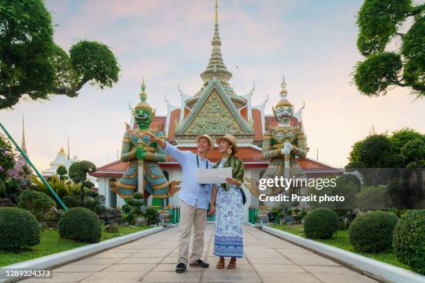 mixed race couple happy tourists to travel on they holidays and holding travel map and pointing in wat arun temple in bangkok, thailand - wat arun tempel stock-fotos und bilder