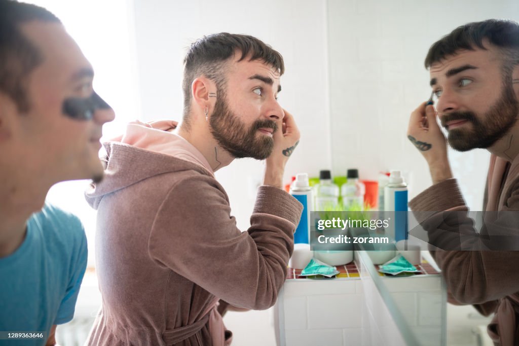 Young man tweezing eyebrows in front of the bathroom mirror with his boyfriend