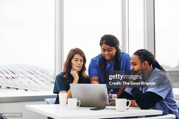 female nurse with laptop talking to colleagues against window in hospital - healthcare leadership stock pictures, royalty-free photos & images