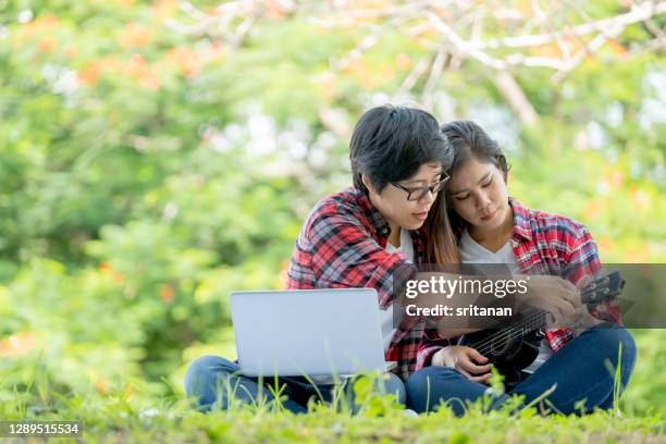couple lgbt women sit in park or garden with one use notebook and teach the other play ukulele or small guitar with morning light - asian lesbians kiss stock pictures, royalty-free photos & images