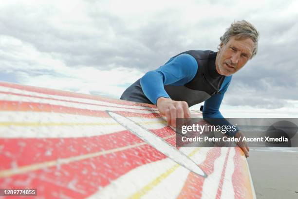 portrait of a mature surfer waxing his board, surf and beach beyond him - wetsuit stock pictures, royalty-free photos & images