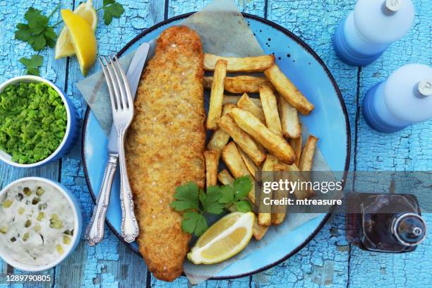 image of fish and chips (cod) garnished with parsley on turquoise plate with greaseproof parchment, knife and fork, lemon slice, bowls containing tartare sauce and mushy peas, bottle of vinegar, turquoise background, elevated view - cod dinner stock pictures, royalty-free photos & images
