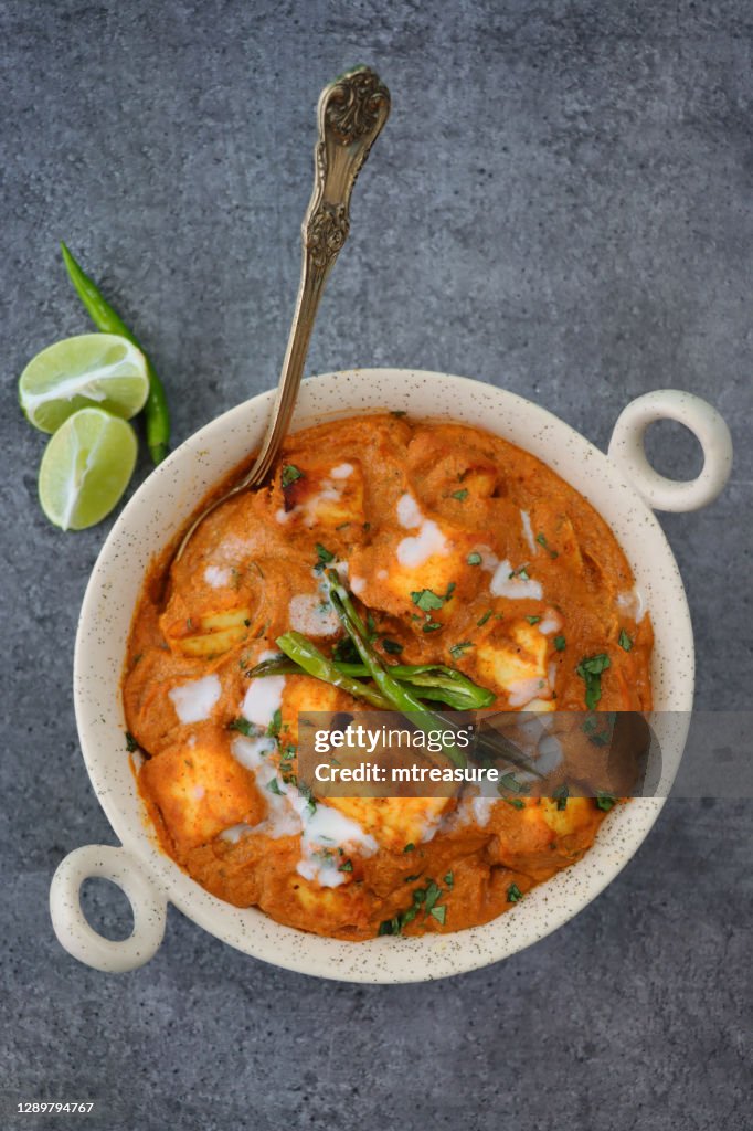 Image of kadhai-style serving bowl filled with homemade Shahi paneer (cheese curry), thick gravy, cream, tomatoes, Indian spices with metal spoon, grey background, elevated view