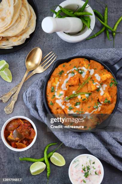 bild von kochpfanne von hausgemachten shahi paneer (käse curry) auf grauem käsetuch, dicke soße, sahne, tomaten, indische gewürze mit lachha paratha (geschichtet es fladenbrot), gabel und löffel, schüssel raita, chutney, mörtel und stößel, erhöht - paneer stock-fotos und bilder