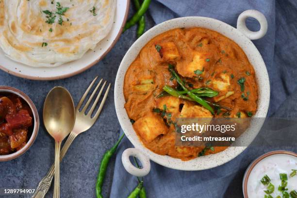 image of kadhai-style serving bowl filled with homemade shahi paneer (cheese curry) on grey cheesecloth, thick gravy, cream, tomatoes, indian spices served with lachha paratha (layered flatbread), fork and spoon, bowls of raita and chutney, elevated view - panir stock pictures, royalty-free photos & images