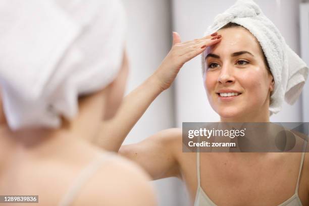 reflection of smiling woman with towel on head rubbing face cream into forehead - face cream stock pictures, royalty-free photos & images