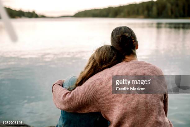 rear view of mother with daughter sitting by lake - women embracing stock pictures, royalty-free photos & images