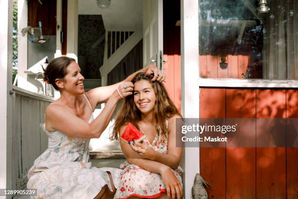 smiling mother tying daughter's hair while sitting outdoors - teenagers eating with mum stock-fotos und bilder