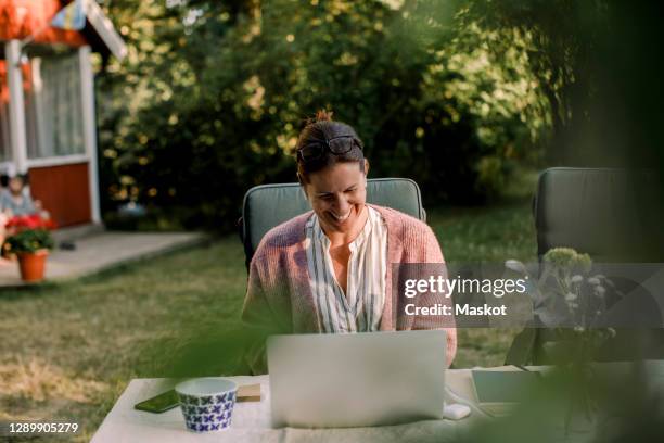 smiling woman using laptop while sitting at dining table in yard during summer - cottage garden stock pictures, royalty-free photos & images