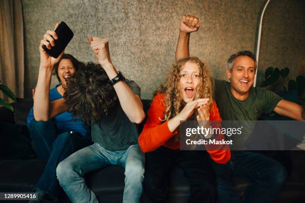 happy parents with children cheering while watching sports in living room at night - family looking at smartphone stockfoto's en -beelden