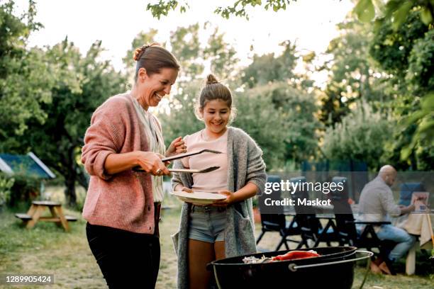 smiling mother and daughter looking at red chili pepper over barbecue grill in yard - familie grillen stock-fotos und bilder