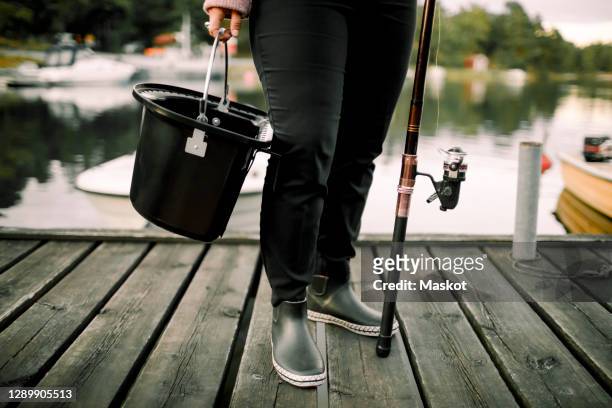 low section of woman with fishing rod and basket standing on pier near lake - wellington boot stockfoto's en -beelden