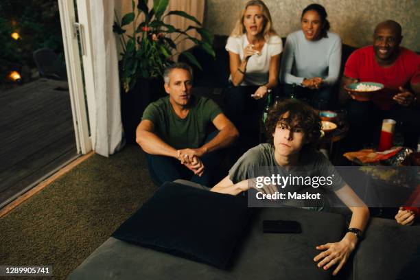 male teenager by sofa watching tv during sporting event with family at night - family game night stock pictures, royalty-free photos & images