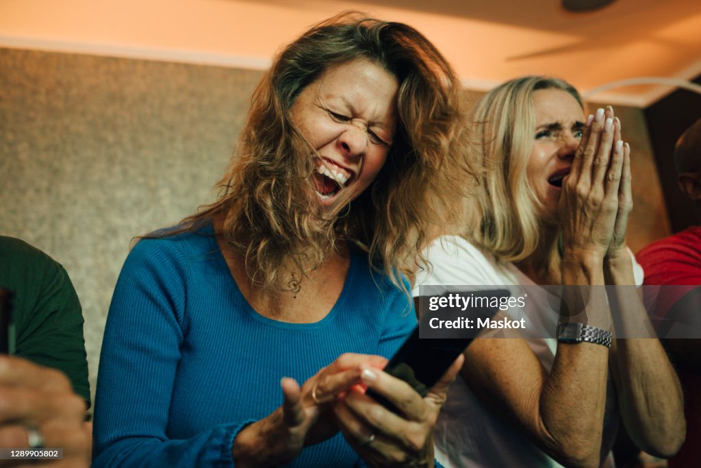 Happy woman cheering by friends while watching sports on smart phone in living room