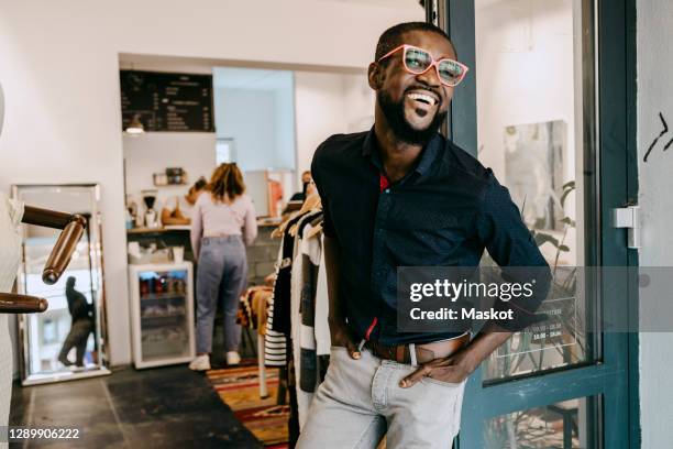 smiling owner with hands in pockets standing at doorway of clothing store - fashion store staff foto e immagini stock