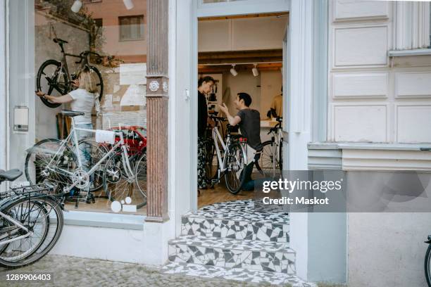 male owner repairing bicycle of senior customer while colleagues working at workshop seen through doorway - bicycle shop 個照片及圖片檔