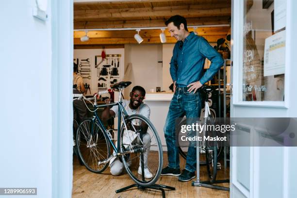 smiling owner repairing bicycle in workshop by colleague seen through glass window - loja de bicicletas imagens e fotografias de stock