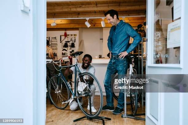 smiling owner repairing bicycle in workshop by colleague seen through glass window - bicycle shop 個照片及圖片檔