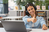 Beautiful happy young woman or student female employee sitting at table in the university library or office with laptop looking at camera and showing thumbs up sign
