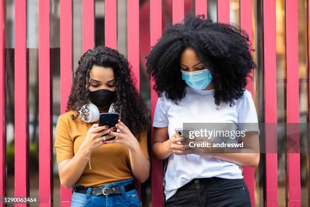 duas mulheres na rua da cidade usando máscara de proteção facial coronavírus e usando o smartphone mantendo a distância social - máscara cirúrgica - fotografias e filmes do acervo