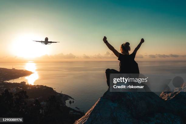 a teenage girl with a backpack sits on the edge of the peak of mountain and watches an airplane flying over the sea coast at sunrise - airplane travel stock-fotos und bilder