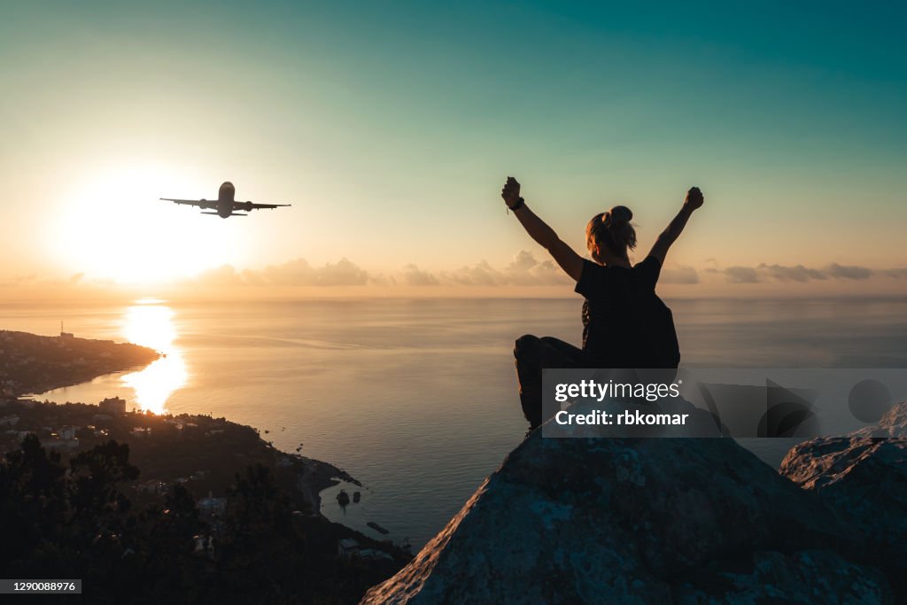 A teenage girl with a backpack sits on the edge of the peak of mountain and watches an airplane flying over the sea coast at sunrise