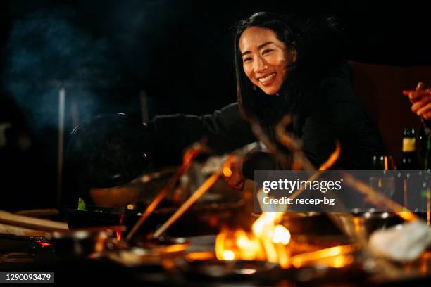 a portrait of young woman among group of friends sitting around camp fire and enjoying food and drink at night in winter - hyper japan stock pictures, royalty-free photos & images