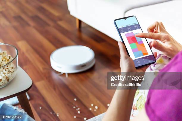 a woman using a robotic vacuum cleaner application on her smartphone - robot vacuum stock pictures, royalty-free photos & images