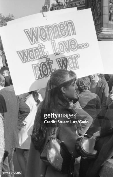 Profile view of a young woman, on a sunny day, holding a sign with the text "Women Want Love..Not War, " participating in Vietnam War protests,...