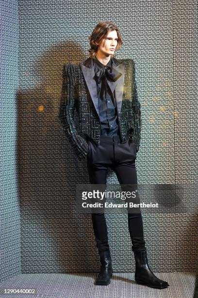 Model wears a black oversized blazer shiny glittering jacket with embroidered Balmain monograms, a bow tie, a black shirt, black suit pants, leather...