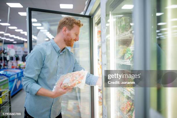 smiling young man taking frozen vegetables from fridge in supermarket - frozen beard stock pictures, royalty-free photos & images