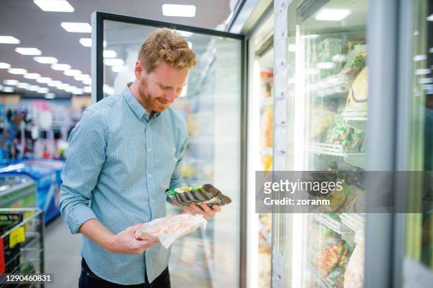 smiling young man choosing in refrigerated section in supermarket - frozen food stock pictures, royalty-free photos & images