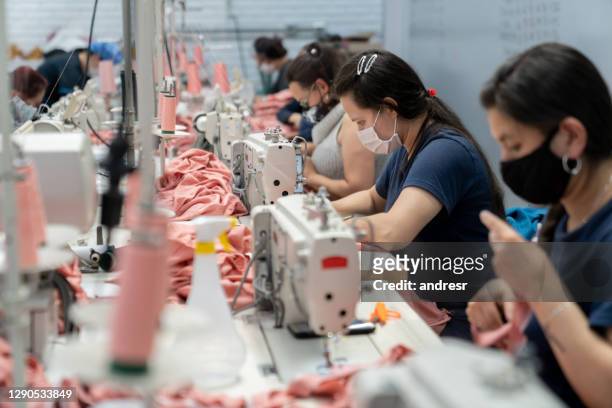 frauen, die in einer textilfabrik arbeiten und eine gesichtsmaske tragen, während sie kleidung nähen - textilfabrik stock-fotos und bilder