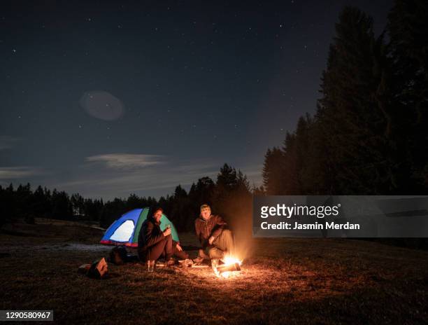 friends with camping tent sitting near forest campfire under starry sky - zelt nacht stock-fotos und bilder