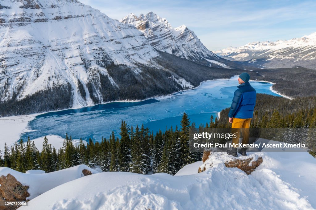 Man stands on snowy knoll above lake and snowcapped mountains