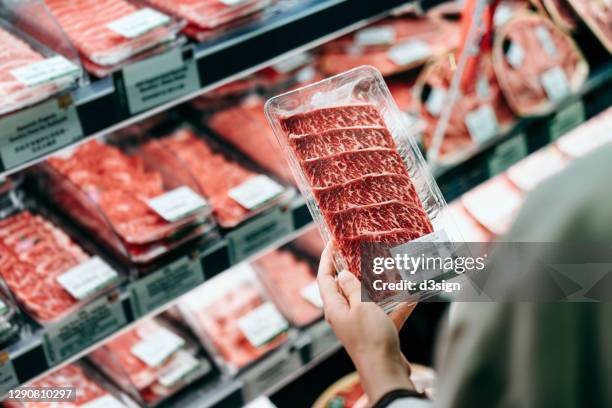 over the shoulder view of young asian woman shopping in a supermarket. she is choosing meat and holding a packet of organic beef in front of the refrigerated section - rood vlees stockfoto's en -beelden