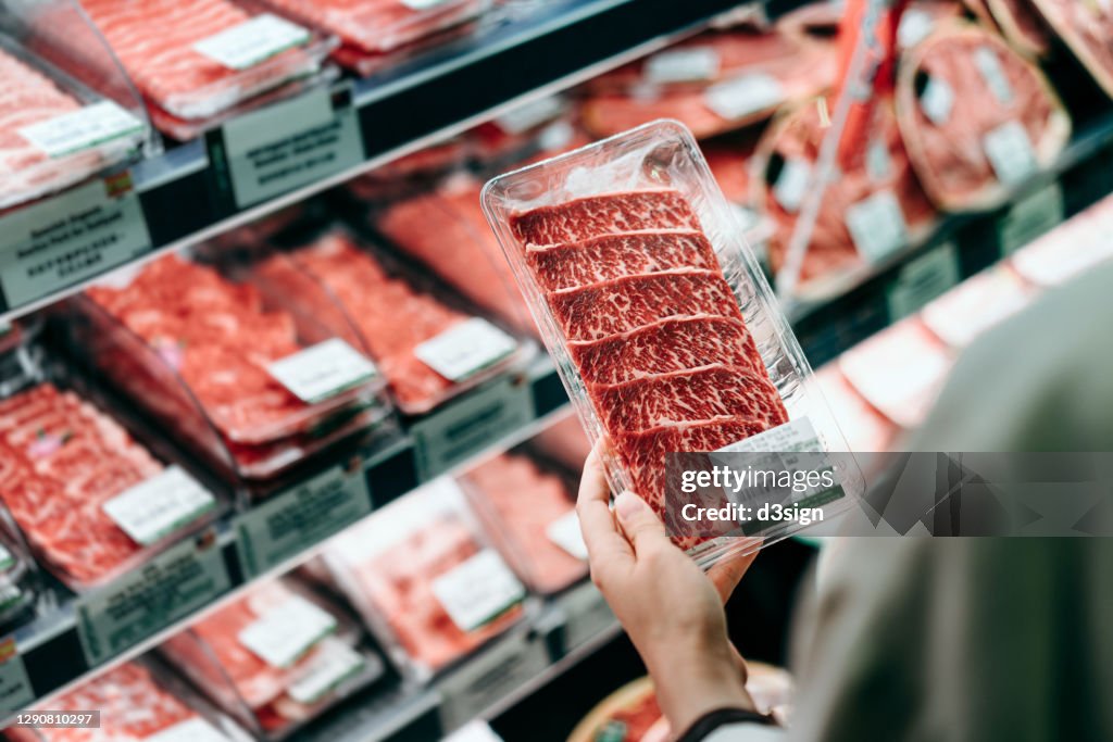 Over the shoulder view of young Asian woman shopping in a supermarket. She is choosing meat and holding a packet of organic beef in front of the refrigerated section